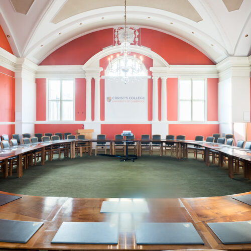 The Plumb Auditorium at Christ’s College, Cambridge, with high ceilings, polished wooden tables and beautiful chandelier lighting. This room is set up in a meeting room style.