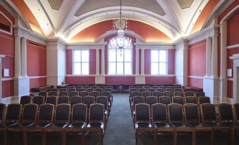 The Plumb Auditorium at Christ’s College, Cambridge, with high ceilings, polished wooden tables and beautiful chandelier lighting. This room is set up in a classroom style.